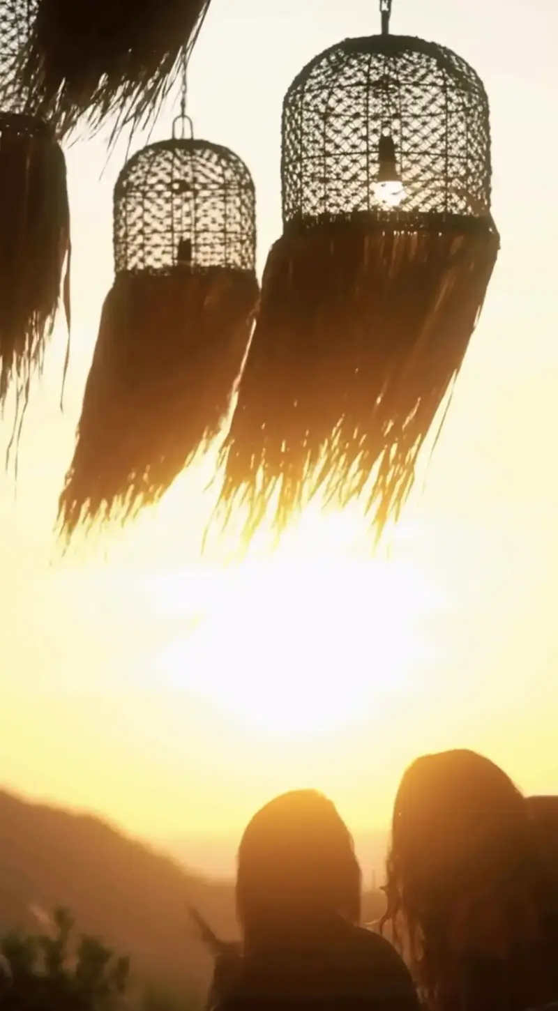 Woven rattan pendant lamps silhouetted against a golden sunset sky above the terrace at Secret Mountain, Koh Phangan, with two guests watching the sun go down over the jungle